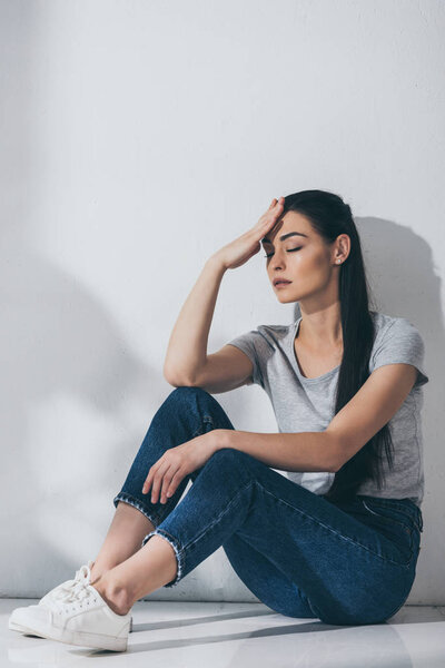 unhappy young woman with closed eyes and hand on forehead sitting on floor near grey wall 