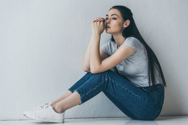 side view of upset young brunette woman sitting and looking away near grey wall