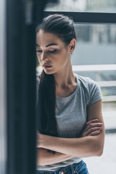 depressive young brunette woman standing with crossed arms near window