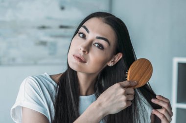 young brunette woman combing hair with hairbrush and looking at camera clipart