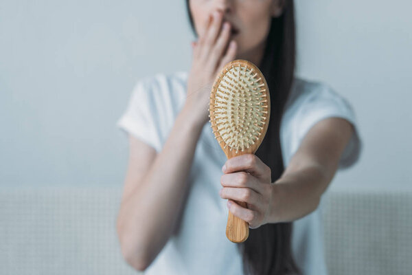 close-up view of shocked young woman holding hairbrush with fallen hair on grey 