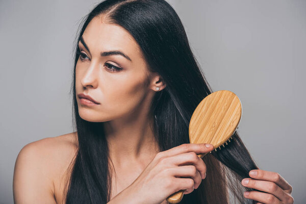 frustrated brunette girl combing hair and looking away isolated on grey