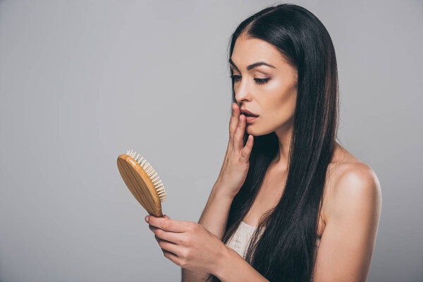 shocked young brunette woman holding hairbrush isolated on grey, hair loss concept 