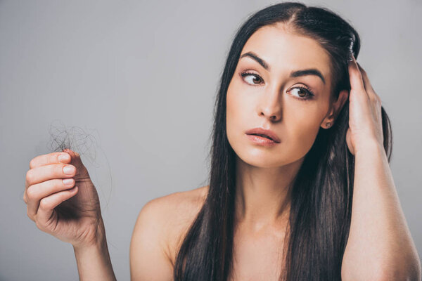 young brunette woman holding fallen hair and looking away isolated on grey, hair loss concept 