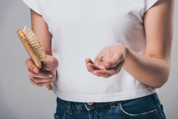 cropped shot of woman holding hairbrush and fallen hair isolated on grey, hair loss concept