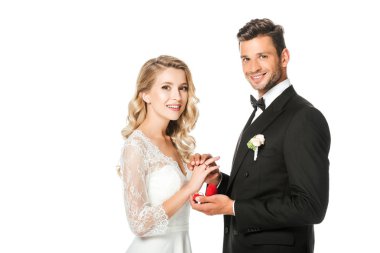happy groom putting on wedding ring on brides finger and looking at camera isolated on white clipart