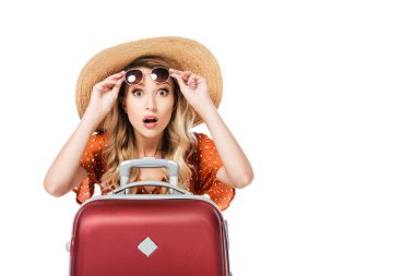 shocked beautiful girl holding sunglasses and sitting near travel bag isolated on white  clipart