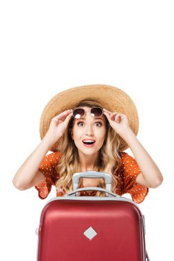 excited beautiful girl holding sunglasses and sitting near travel bag isolated on white  clipart