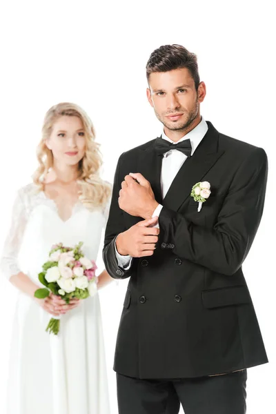 英俊的年轻新郎扣袖扣能够和看着在相机与新娘站模糊的背景上孤立的白色 — 图库照片