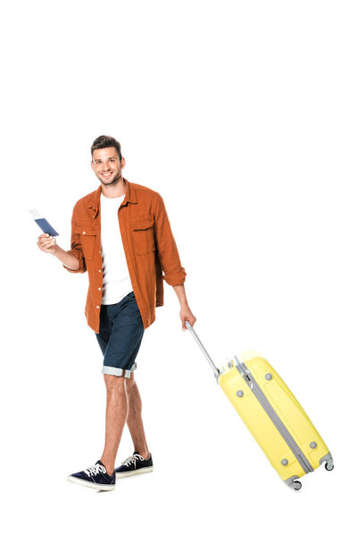 happy young man with luggage and flight ticket walking and looking at camera isolated on white