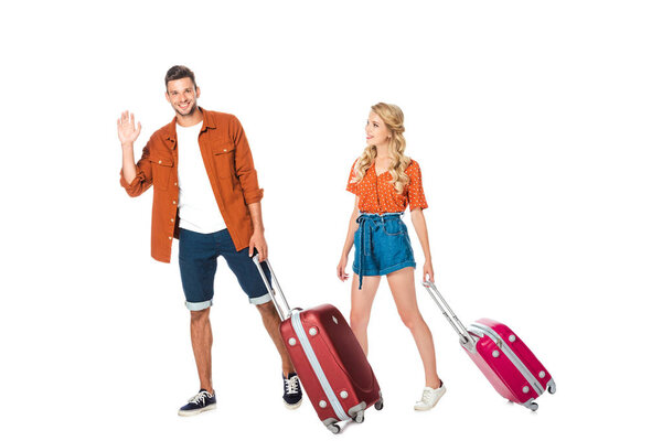 happy young couple with luggage walking and waving at camera isolated on white