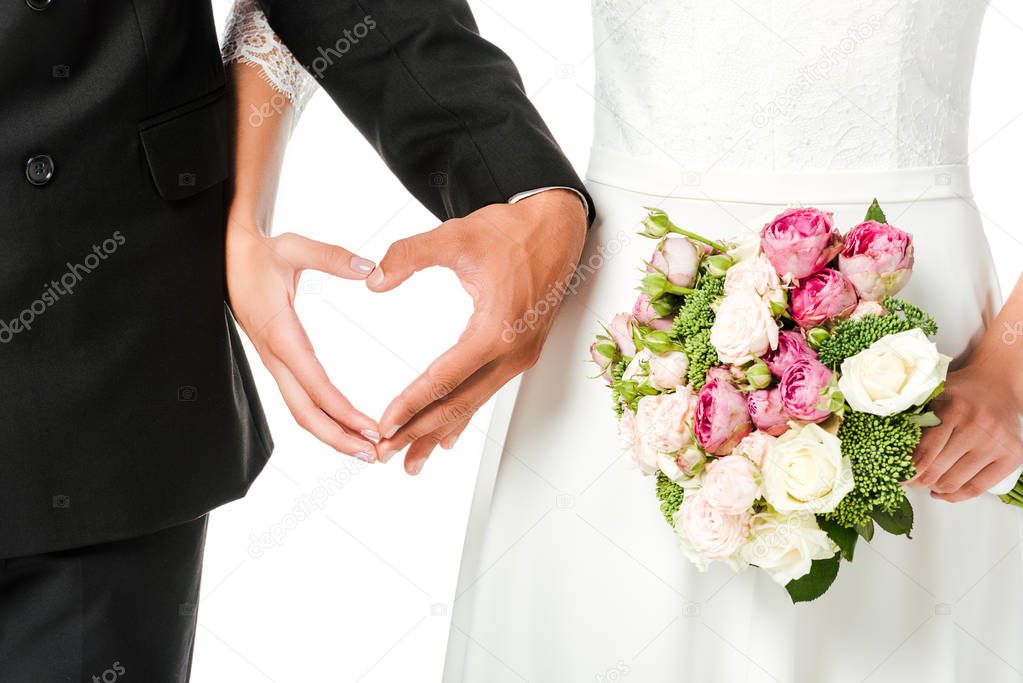 cropped shot of bride with bouquet and groom making heart sign with hands isolated on white