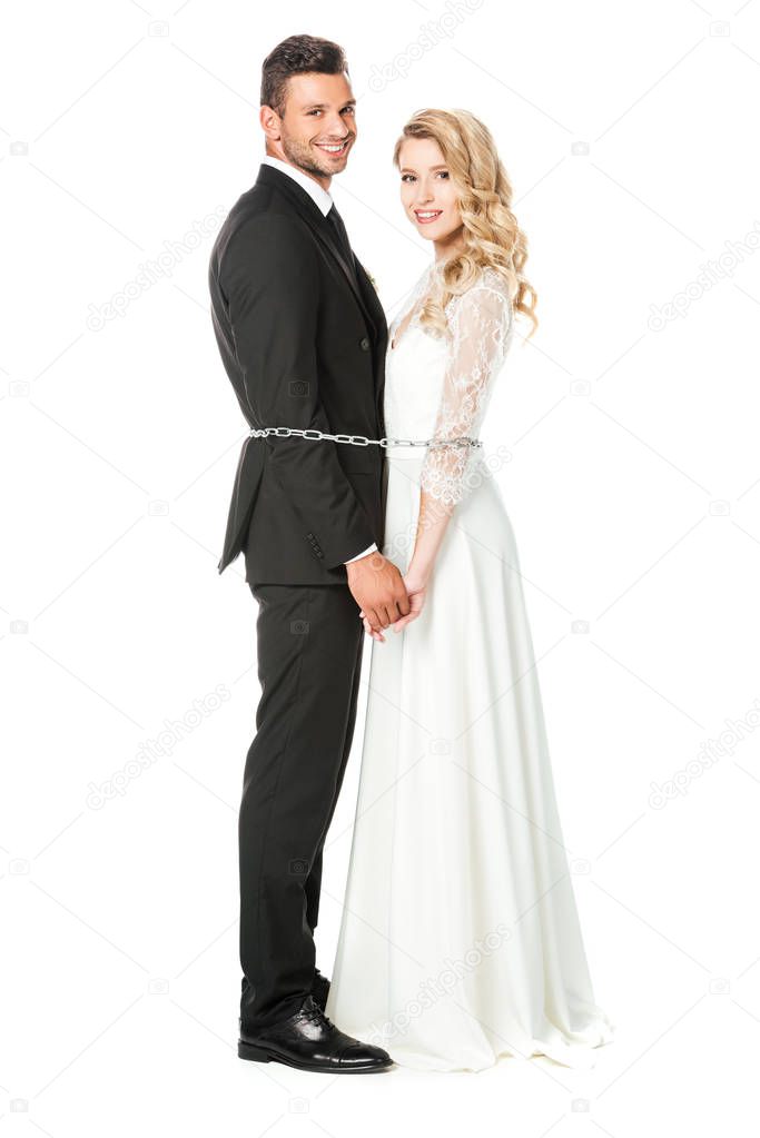 happy young newlyweds tied with chain back to back and looking at camera isolated on white