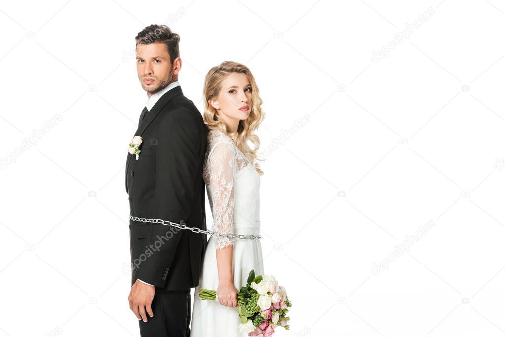 young newlyweds tied with chain back to back and looking at camera isolated on white