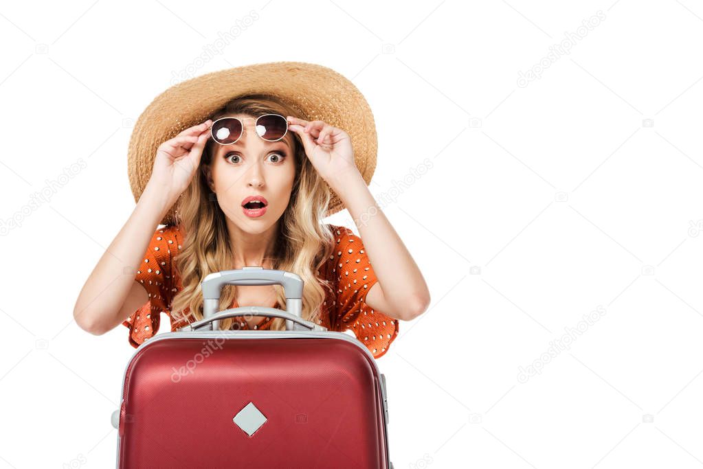 shocked beautiful girl holding sunglasses and sitting near travel bag isolated on white 