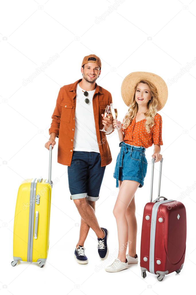 smiling couple standing with travel bags and clinking with glasses of champagne isolated on white