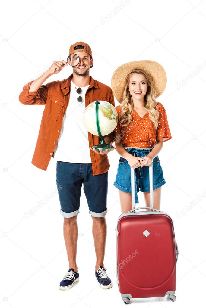 smiling couple standing with magnifying glass, globe and travel bag isolated on white