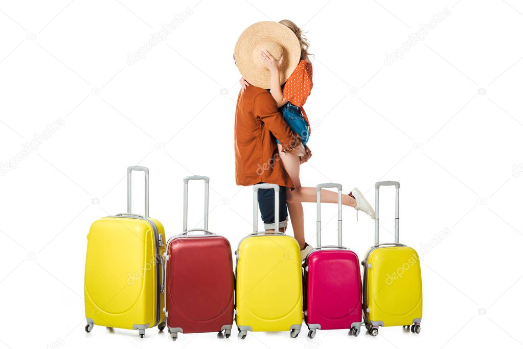 obscured view of newlywed couple kissing behind straw hat with arranged suitcases around isolated on white