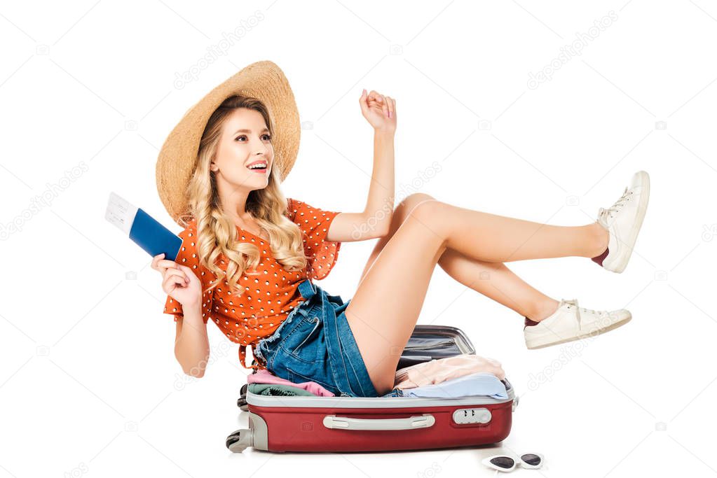 happy young woman in straw hat with passport and ticket sitting in suitcase isolated on white