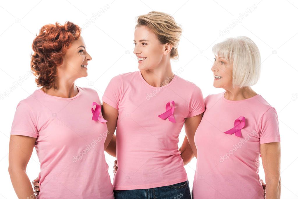 women with pink ribbons standing together and smiling each other isolated on white, breast cancer awareness concept