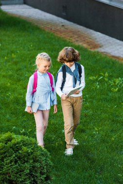 high angle view of schoolkids with backpacks holding digital tablet while standing together on green lawn  clipart