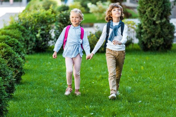 Вид в полный рост восхитительных счастливых школьников с рюкзаками, держащихся за руки и идущих по зеленому полотну
