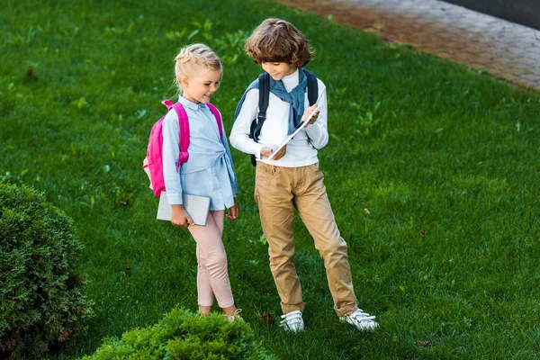 緑の芝生の上に立っている間デジタル タブレットを一緒に保持するバックパックと子供観高角度  — 無料ストックフォト