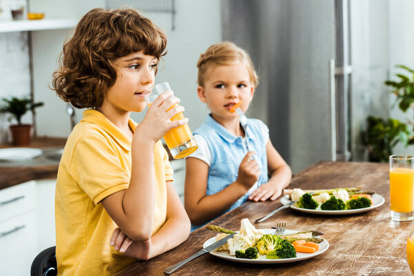 adorable kids sitting together at table, eating vegetables and drinking juice