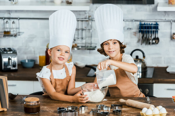 cute little children in chef hats preparing dough for cookies and smiling at camera