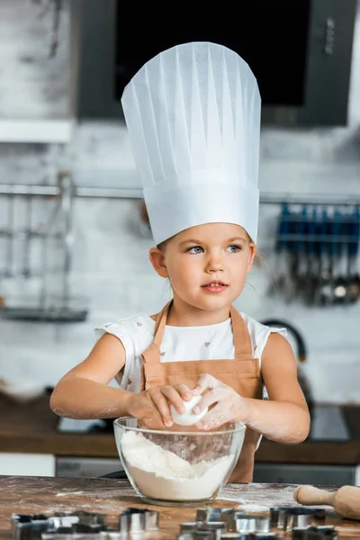 adorable child in aron and chef hat cooking dough and looking away in kitchen