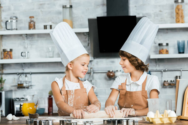 adorable happy kids in chef hats preparing dough for cookies and smiling each other