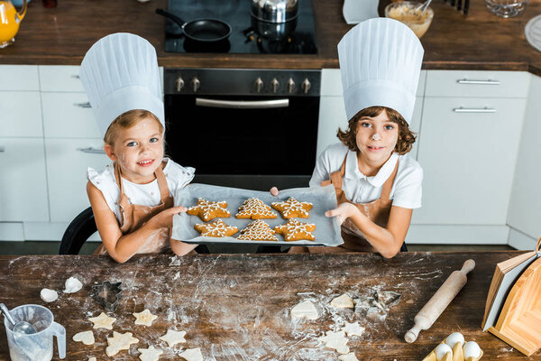 high angle view of happy kids in chef hats holding baking tray with ginger cookies and smiling at camera