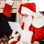 Focused santa claus using tablet while sitting in armchair near fireplace isolated on white