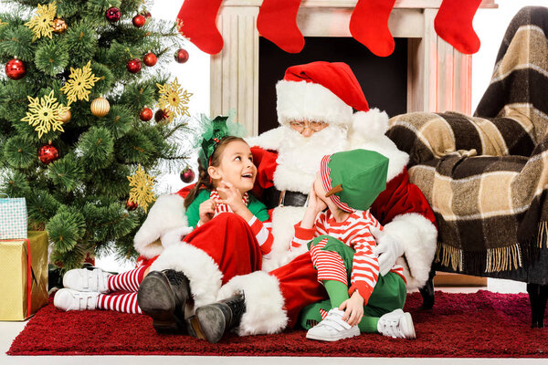 santa embracing happy kids while they sitting on floor near fireplace together