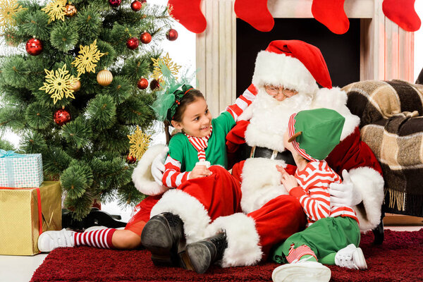 santa embracing excited kids while they sitting on floor in front of fireplace together