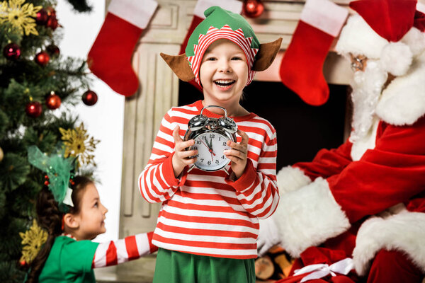adorable little kid holding alarm clock with his sister and santa passing gift box on background