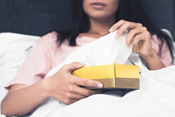 cropped shot of sick young woman taking paper napkin out of box while sitting in bed