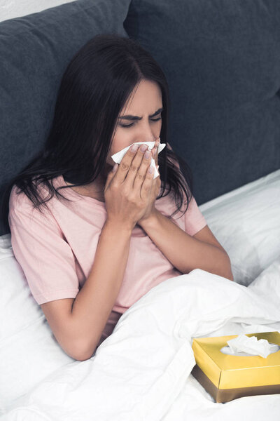 sick young woman with box of paper napkins sneezing while sitting in bed