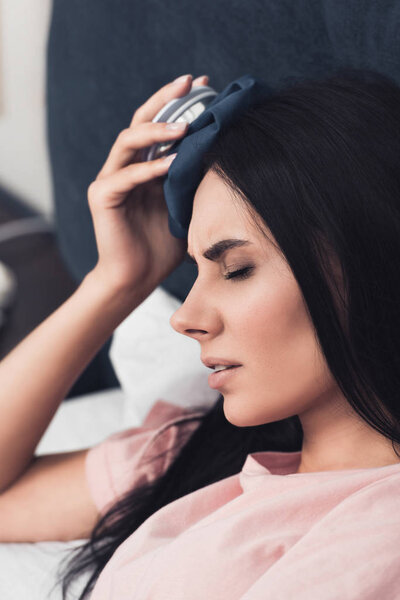 close-up shot of sick young woman holding ice pack on head while lying in bed