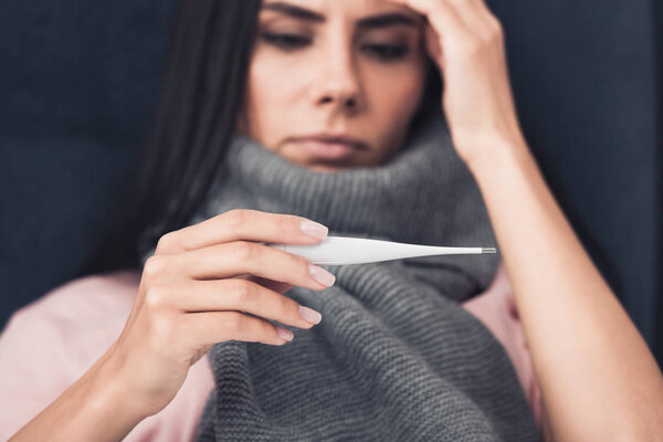 close-up shot of sick young woman in scarf looking at electronic thermometer
