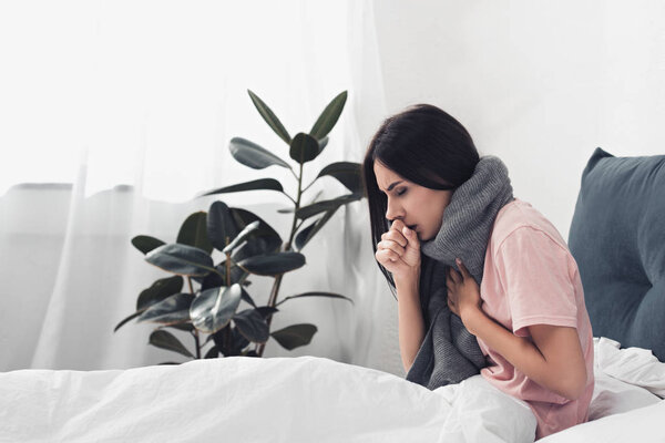 больная молодая женщина сидит в постели и кашляет, страдая от боли в горле
