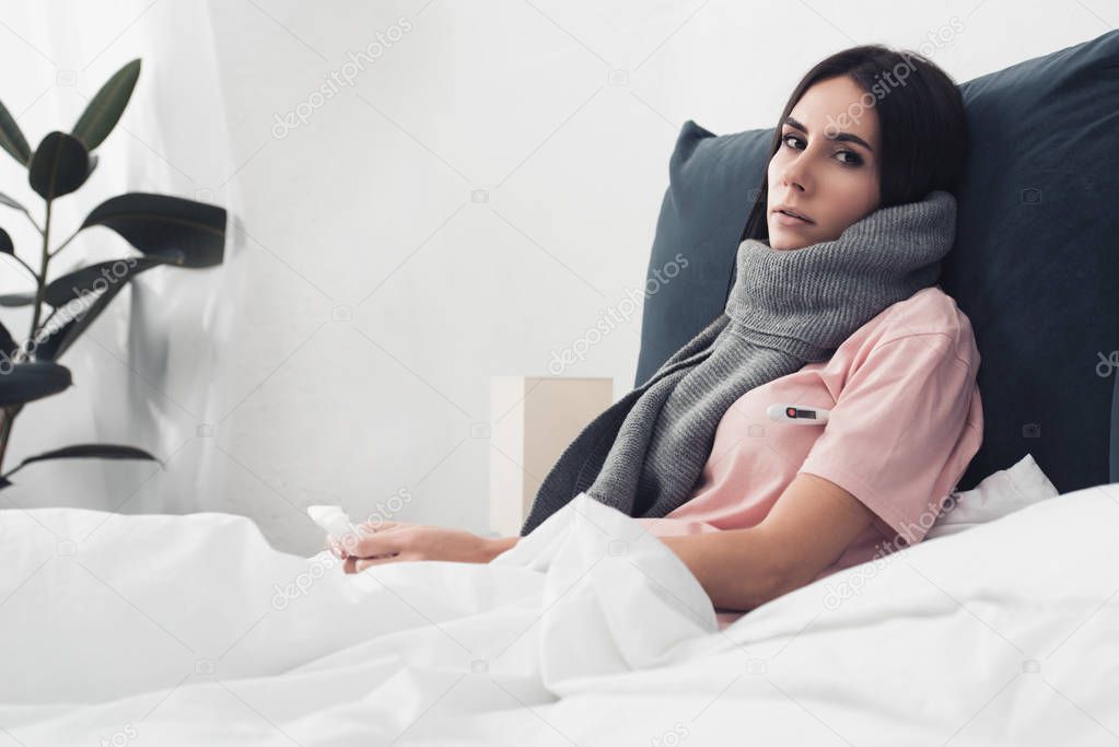 exhausted sick young woman in scarf sitting in bed and looking at camera