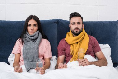 serious sick couple with pills and glasses of water sitting in bed and looking at camera clipart