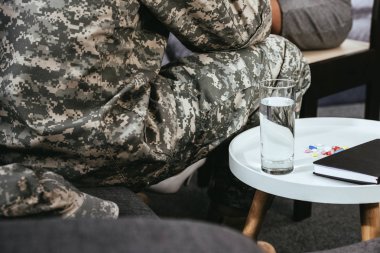 cropped shot of soldier sitting on couch with glass of water and pills on table