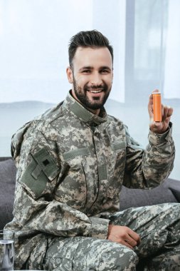 happy young soldier holding plastic jar of pills and looking at camera clipart