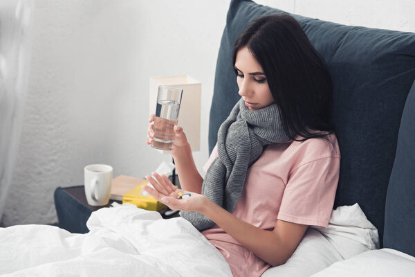 diseased young woman holding pills and glass of water while lying in bed