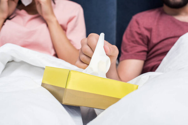 обрезанный снимок больной пары, сидящей в постели и вынимающей бумажные салфетки из коробки
