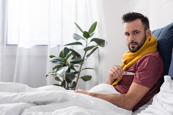 diseased young man measuring temperature with electronic thermometer in bed