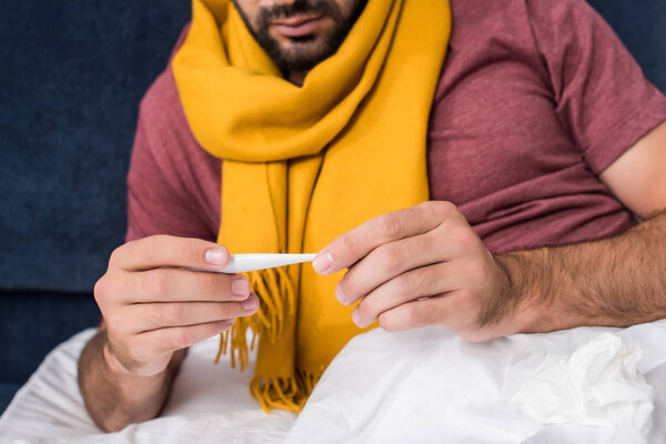 cropped shot of sick young man holding electronic thermometer while lying in bed