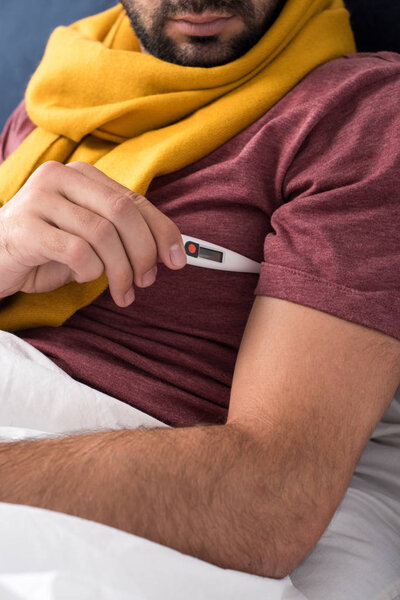 cropped shot of sick man measuring temperature with electronic thermometer in bed